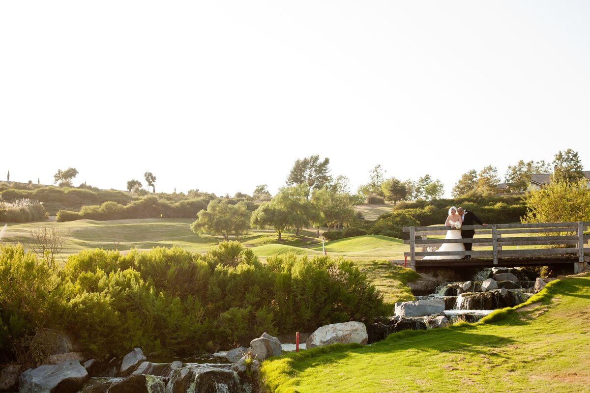 A bride and groom on a wooden footbridge above a small waterfall at a golf-course venue in late-afternoon light